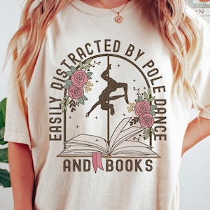 May include: A white t-shirt with the text "Easily Distracted by Pole Dance and Books" in brown lettering. The text is surrounded by a circle with a silhouette of a woman pole dancing and pink flowers.