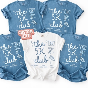 May include: Five t-shirts, four in a blue hue and one white, featuring the text "the 5K club" in a handwritten font. The design includes a 5K badge, a medal, and a running shoe. The text "Portland, OR" and "Team Name Here" are also present.