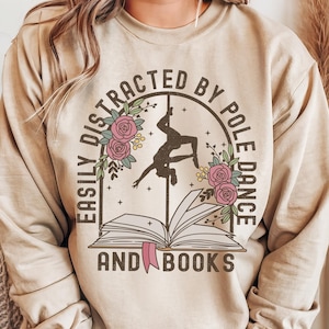 May include: Sand-coloured sweatshirt with the text "EASILY DISTRACTED BY POLE DANCE AND BOOKS". The design features a silhouette of a pole dancer, flowers, and an open book. The sweatshirt is a neutral colour.
