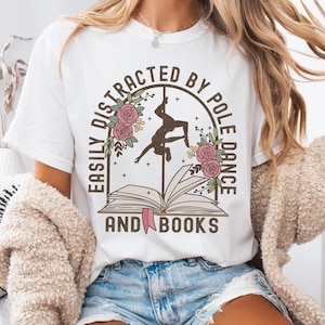 Puede incluir: Camiseta blanca con un diseño gráfico de un libro con una bailarina de pole dance dentro. El texto dice "Easily Distracted by Pole Dance and Books".