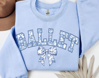 Puede incluir: Una sudadera azul claro con la palabra "BALLET" en un estampado floral. Un lazo a juego está debajo de la palabra. La sudadera está doblada sobre una superficie blanca con un sombrero marrón y una planta seca en el fondo.