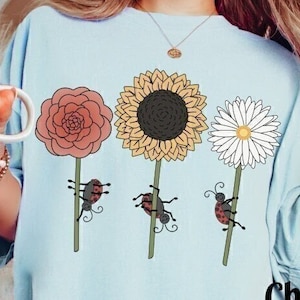 May include: A light blue t-shirt with a graphic of three flowers with ladybugs climbing up the stems. The flowers are a red rose, a yellow sunflower, and a white daisy. The text " Chambray" is printed on the bottom right corner of the shirt.