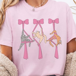 May include: A pink t-shirt with a graphic of three cats hanging from pink bows. The cats are in different poses and have different colored fur.
