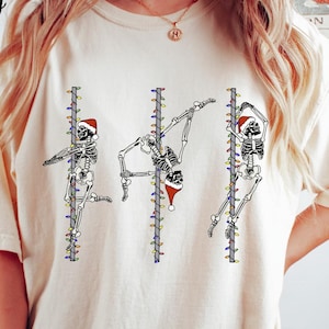 May include: Ivory-coloured t-shirt featuring three skeleton figures pole dancing, each wearing a Santa hat and surrounded by Christmas lights. The word "Ivory" is printed in the upper left corner.