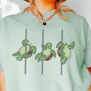 May include: A light green t-shirt featuring three cartoon turtles on poles. The turtles are green with brown shells and are in various poses. The word "Bay" is printed in black at the bottom left.