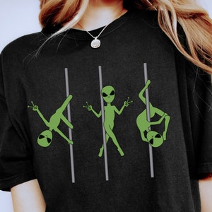 Könnte beinhalten: Schwarzes T-Shirt mit einem Aufdruck von drei grünen Außerirdischen, die an einer Stange tanzen. Die Außerirdischen halten jeweils ein Peace-Zeichen.