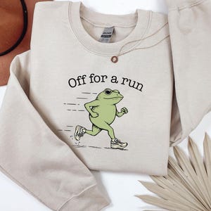 Puede incluir: Sudadera de color arena con el texto "Off for a run" y una rana de dibujos animados con zapatillas deportivas. La rana está corriendo. La sudadera tiene mangas largas y cuello redondo.