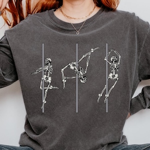 May include: A dark grey long-sleeved sweatshirt featuring a graphic of three skeletons dancing on a pole. The model is wearing blue jeans and white trainers.