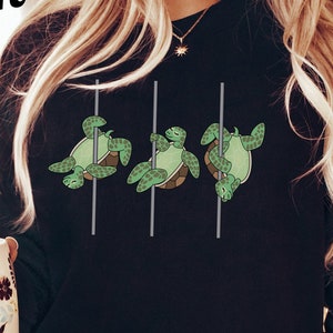 May include: Black sweatshirt with a graphic of three turtles on a pole dance. The turtles are green and the poles are gray. The sweatshirt is being worn by a person with long blonde hair and blue jeans.