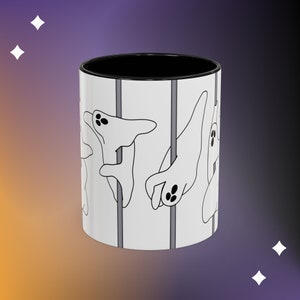 May include: White ceramic mug with a black rim and handle. The mug features a design of ghosts hanging from poles.