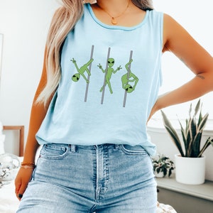 Op de afbeelding: Een lichtblauw tanktop met een afbeelding van drie groene aliens die aan het pole-dansen zijn. Het tanktop wordt gedragen door een persoon met lang blond haar en een blauwe jeans.