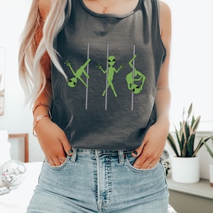 Op de afbeelding: Een grijs tanktop met een afbeelding van drie groene aliens die een paaldans uitvoeren. Het tanktop wordt gedragen door een persoon met lang blond haar en een blauwe jeans.