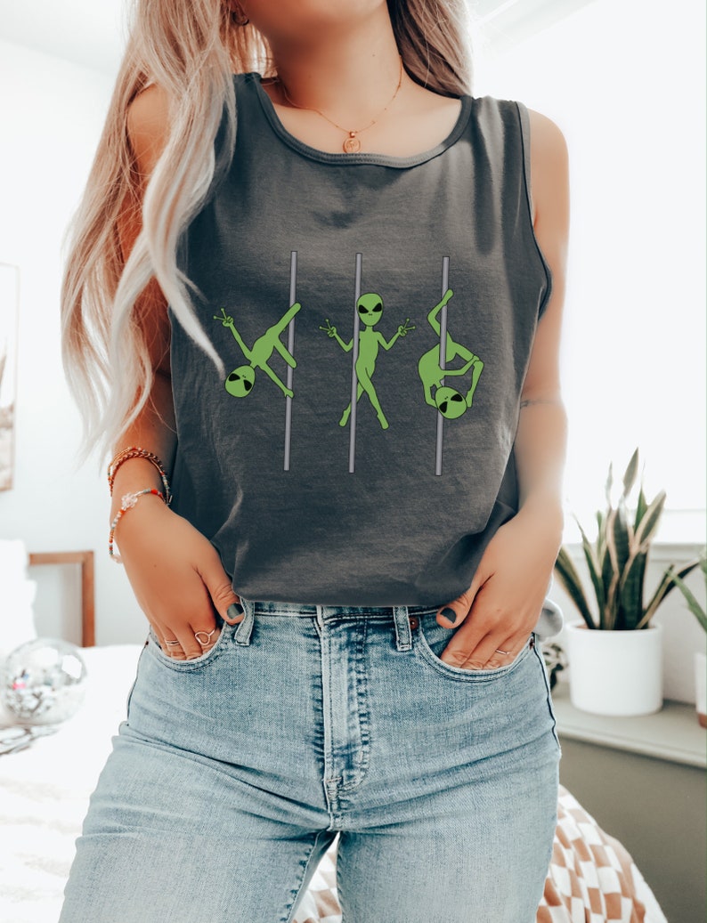 Op de afbeelding: Een grijs tanktop met een afbeelding van drie groene aliens die een paaldans uitvoeren. Het tanktop wordt gedragen door een persoon met lang blond haar en een blauwe jeans.