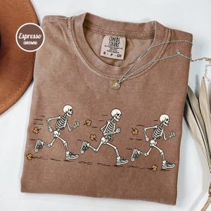May include: An espresso brown crewneck t-shirt featuring a graphic of three skeletons running across the front, accompanied by scattered autumn leaves. The inner neck label reads Comfort Colors, and a silver chain necklace rests on the collar.