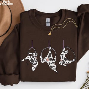 May include: A dark chocolate brown sweatshirt with a design of three cartoon cows doing aerial acrobatics on hoops. The cows are white with black spots. A gold star pendant necklace is on the sweatshirt. The words "Dark Chocolate" are in the upper left corner.