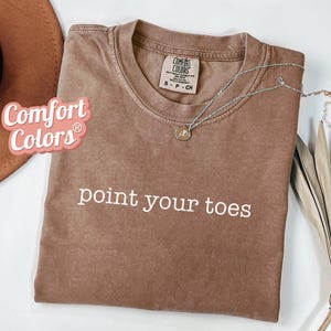 May include: A brown Comfort Colors t-shirt with the phrase "point your toes" in white. A silver necklace with a small pendant rests on the shirt. A brown hat and dried plant are in the background.