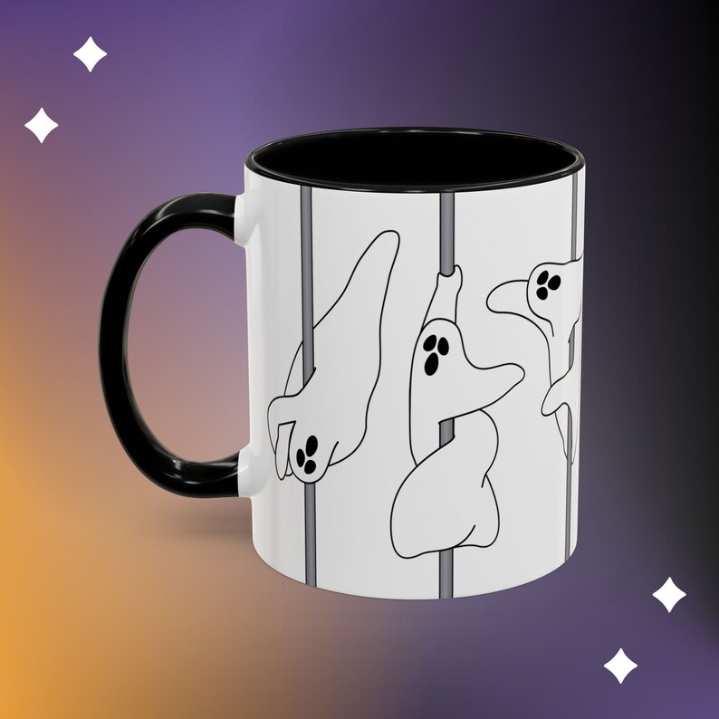 May include: A white ceramic coffee mug with a black handle and rim. The mug features a black and white illustration of three ghosts hanging from a pole.