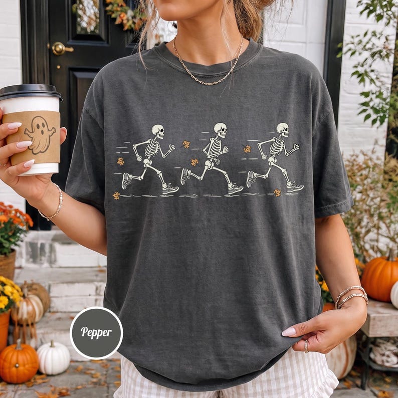 May include: A charcoal gray short-sleeve t-shirt featuring a graphic print of three white skeletons running in a row, interspersed with small orange autumn leaves. The shirt has a crew neckline and a relaxed fit, with the word Pepper printed below.