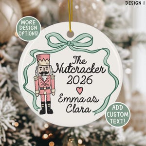 Puede incluir: Adorno navideño de cerámica blanca con una ilustración de cascanueces rosa y negro. El adorno tiene el texto "The Nutcracker 2026" y "Emma as Clara". Un diseño de cinta verde rodea el texto. Incluye el texto "More Design Options!" y "Add Custom Text!"