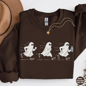 May include: A dark brown crewneck sweatshirt featuring a graphic of three white ghosts running in sequence. The ghosts wear sneakers, and the third ghost holds a water bottle. A gold-toned star pendant necklace rests on the neckline of the garment.