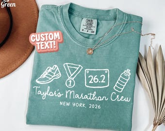 May include: A light green t-shirt with white graphics of a running shoe, medal, race bib with "26.2", and water bottle. The text "Taylor's Marathon Crew NEW YORK, 2026" is printed below the graphics. The words "CUSTOM TEXT!" are in a pink bubble.