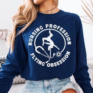 Puede incluir: Sudadera azul marino de manga larga con un gráfico blanco. El gráfico presenta a una bailarina de barra dentro de un círculo con las palabras "Nursing Profession" y "Flying Obsession" y un estetoscopio.