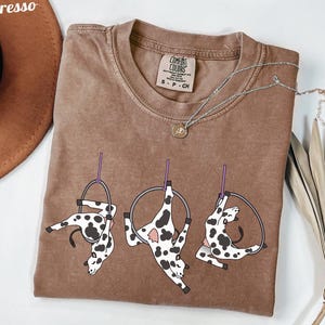 Può includere: Una maglietta marrone con una grafica di tre mucche che eseguono acrobazie aeree su cerchi. Le mucche sono bianche con macchie nere. La maglietta ha un girocollo e maniche corte. Una collana d'argento con un piccolo ciondolo è sulla maglietta.
