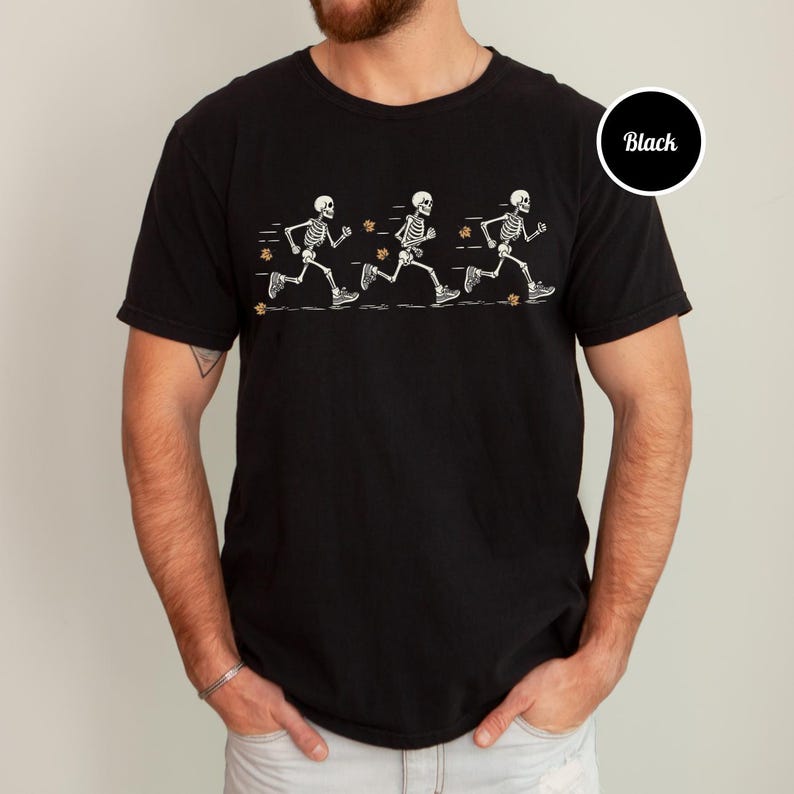 May include: A black short-sleeved t-shirt featuring a graphic print of three white skeletons in motion, depicted in a running sequence. The design includes small orange autumn leaves scattered around the skeletons and horizontal motion lines.