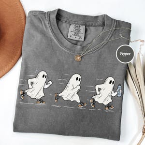 May include: A charcoal gray t-shirt featuring a graphic of three white ghosts running in sequence. The ghosts are wearing orange and black sneakers, and the third ghost is holding a water bottle. The shirt has a Comfort Colors brand neck label.