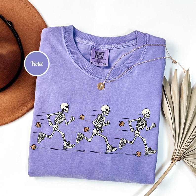May include: A violet short-sleeved t-shirt featuring a graphic of three white skeletons running in a line, accompanied by small orange autumn leaves. The shirt has a Comfort Colors label at the inner neckline and a thin gold-toned chain necklace.