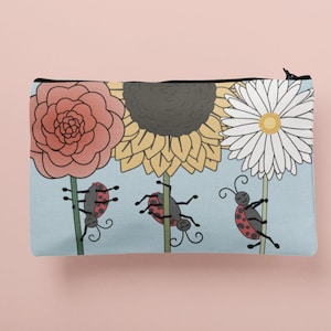 Könnte beinhalten: Eine blaue Reißverschlusstasche mit einem Blumendesign. Drei rote Marienkäfer klettern an den Stielen der Blumen hoch. Die Blumen sind eine rosa Rose, eine gelbe Sonnenblume und eine weiße Gänseblümchen.