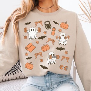 May include: A beige crewneck sweatshirt featuring a repeating pattern of white ghosts lifting orange barbells and dumbbells, interspersed with orange pumpkins, brown bats, orange bows, a yoga mat, a kettlebell, a water bottle, and autumn leaves.