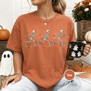May include: A burnt orange short-sleeve t-shirt featuring a graphic of three white skeletons running in a sequence. The skeletons are depicted in motion with scattered brown autumn leaves around them. The shirt has a crew neckline and a relaxed fit.