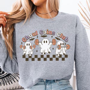 May include: A heather grey crewneck sweatshirt featuring a graphic print of three white ghosts lifting orange dumbbells. The text "Ghoul Gains Club" is arched above the ghosts, accompanied by small black bats and stars, all set above a black and brown checkerboard pattern.