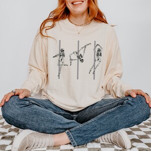 May include: A cream-coloured long-sleeved shirt featuring a graphic of three skeletons performing a pole dance. The shirt is being worn by a person sitting on a chequered floor.