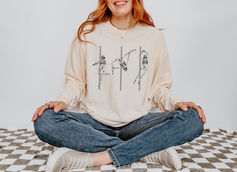 May include: A cream-coloured long-sleeved shirt featuring a graphic of three skeletons performing a pole dance. The shirt is being worn by a person sitting on a chequered floor.