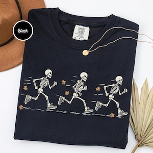 May include: A black crew neck t-shirt featuring a graphic of three white skeletons running in a line. The skeletons are depicted in motion with motion lines and scattered orange autumn leaves. The shirt has a Comfort Colors brand label inside.