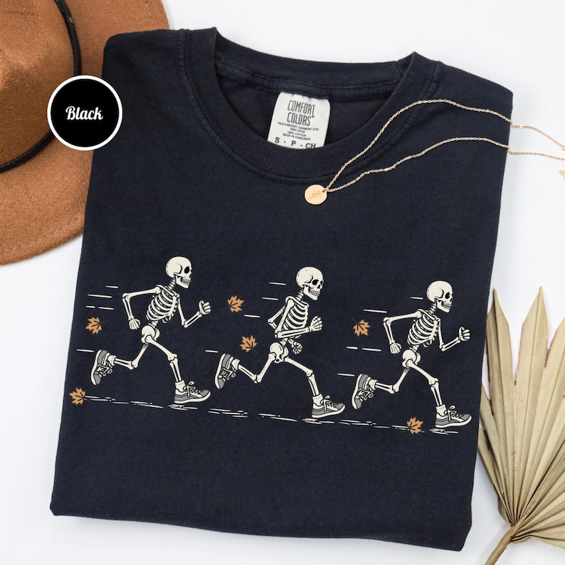 May include: A black crew neck t-shirt featuring a graphic of three white skeletons running in a line. The skeletons are depicted in motion with motion lines and scattered orange autumn leaves. The shirt has a Comfort Colors brand label inside.