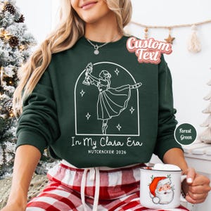 May include: A forest green crewneck sweatshirt featuring a white line art graphic of a ballerina holding a nutcracker inside an arched frame. The text reads In My Clara Era and NUTCRACKER 2026. The sweatshirt is paired with red and white plaid trousers.