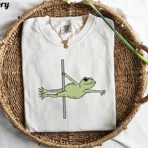 May include: Ivory-colored t-shirt featuring a cartoon frog doing a pole dance. The frog is light green with black eyes and is holding onto a gray pole. The word "Ivory" is printed in black at the top left.
