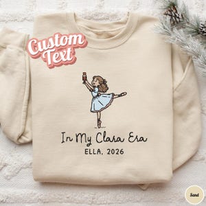 May include: A sand-coloured crewneck sweatshirt featuring an embroidered illustration of a ballerina in a light blue dress holding a nutcracker. Below the figure, the text reads "In My Clara Era" followed by "ELLA, 2026" in black lettering.