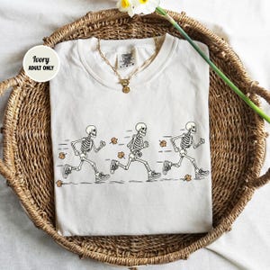 May include: An ivory t-shirt featuring a graphic of three skeletons running in a line, accompanied by small orange autumn leaves. The shirt has a crew neckline and a gold-toned chain necklace resting on the collar. A circular tag reads Ivory Adult Only.