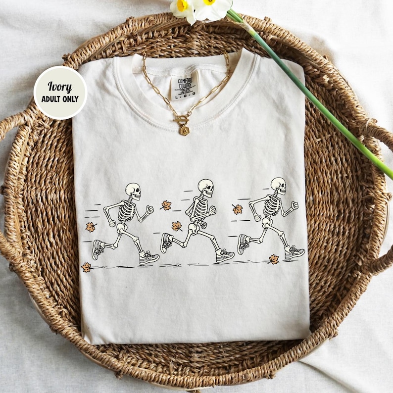 May include: An ivory t-shirt featuring a graphic of three skeletons running in a line, accompanied by small orange autumn leaves. The shirt has a crew neckline and a gold-toned chain necklace resting on the collar. A circular tag reads Ivory Adult Only.