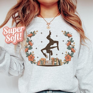 May include: A light grey sweatshirt featuring a floral wreath design and a silhouette of a person pole dancing on a stack of books. The text "Super Soft!" is on the left side of the sweatshirt.