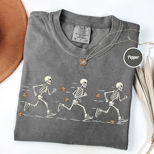 May include: A charcoal gray t-shirt featuring a graphic of three white skeletons running, accompanied by scattered orange autumn leaves. The shirt has a crew neckline and a Comfort Colors brand label on the inside collar with the text Pepper.