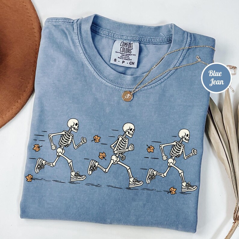 May include: A blue Comfort Colors t-shirt featuring a graphic of three skeletons running across the front, surrounded by scattered orange autumn leaves. The shirt has a crew neckline and a tag that reads Comfort Colors S P CH.