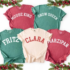 May include: Five short-sleeved t-shirts in various colors: red, green, and cream. Each shirt features a different name in a bold, arched font: "MOUSE KING", "SNOW QUEEN", "FRITZ", "CLARA", and "MARZIPAN". Festive holiday theme.
