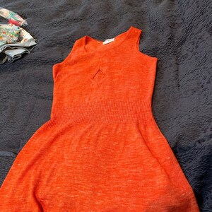 May include: A sleeveless, orange, knit dress with a small diamond-shaped cutout in the front. The dress has a fitted bodice and a flared skirt.