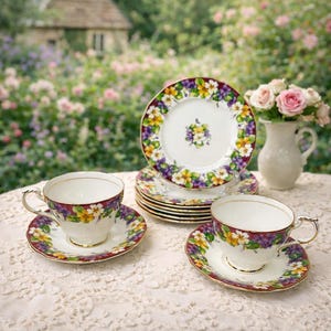 May include: A vintage tea set featuring floral designs in yellow, purple, and white on a burgundy rim. The set includes teacups, saucers, and plates, arranged on a lace tablecloth. A vase of pink roses is also present.