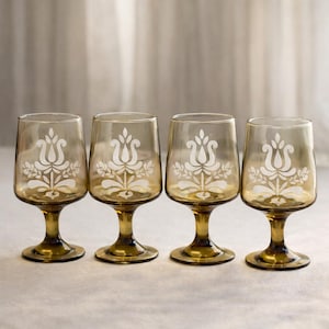 May include: Set of four amber-colored stemmed glasses. Each glass features a white floral design. The glasses have a rounded bowl shape and a short stem. Ideal for serving beverages, these glasses add a touch of vintage charm to any table setting.